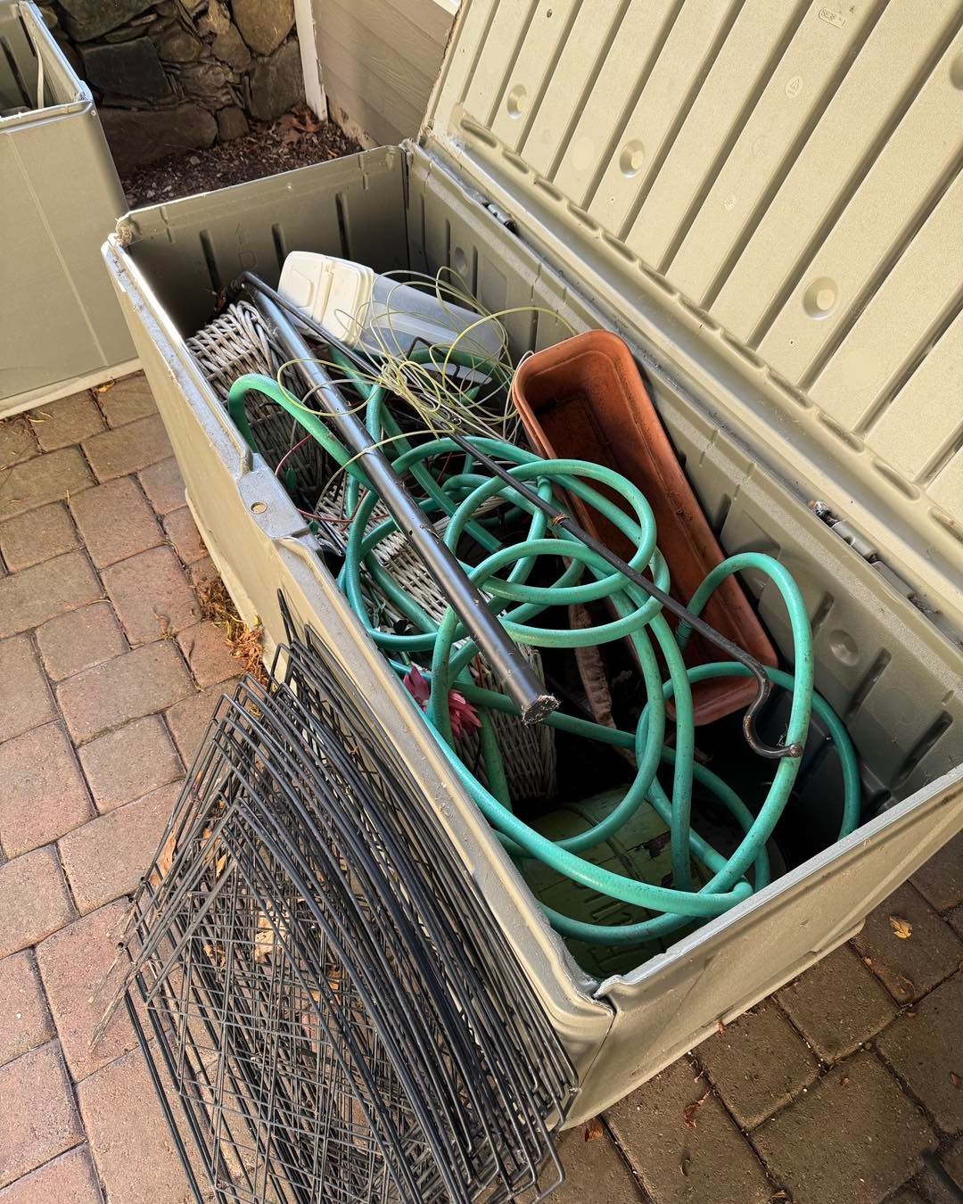 Open storage bin filled with green hoses, black wire mesh, and assorted yard tools outdoors