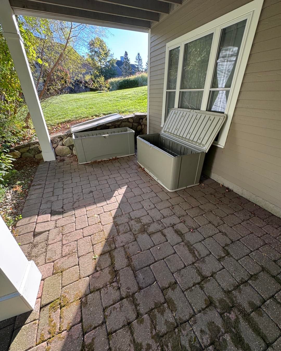 Covered patio with brick pavers, beige siding, windows, and two storage benches overlooking a grassy yard
