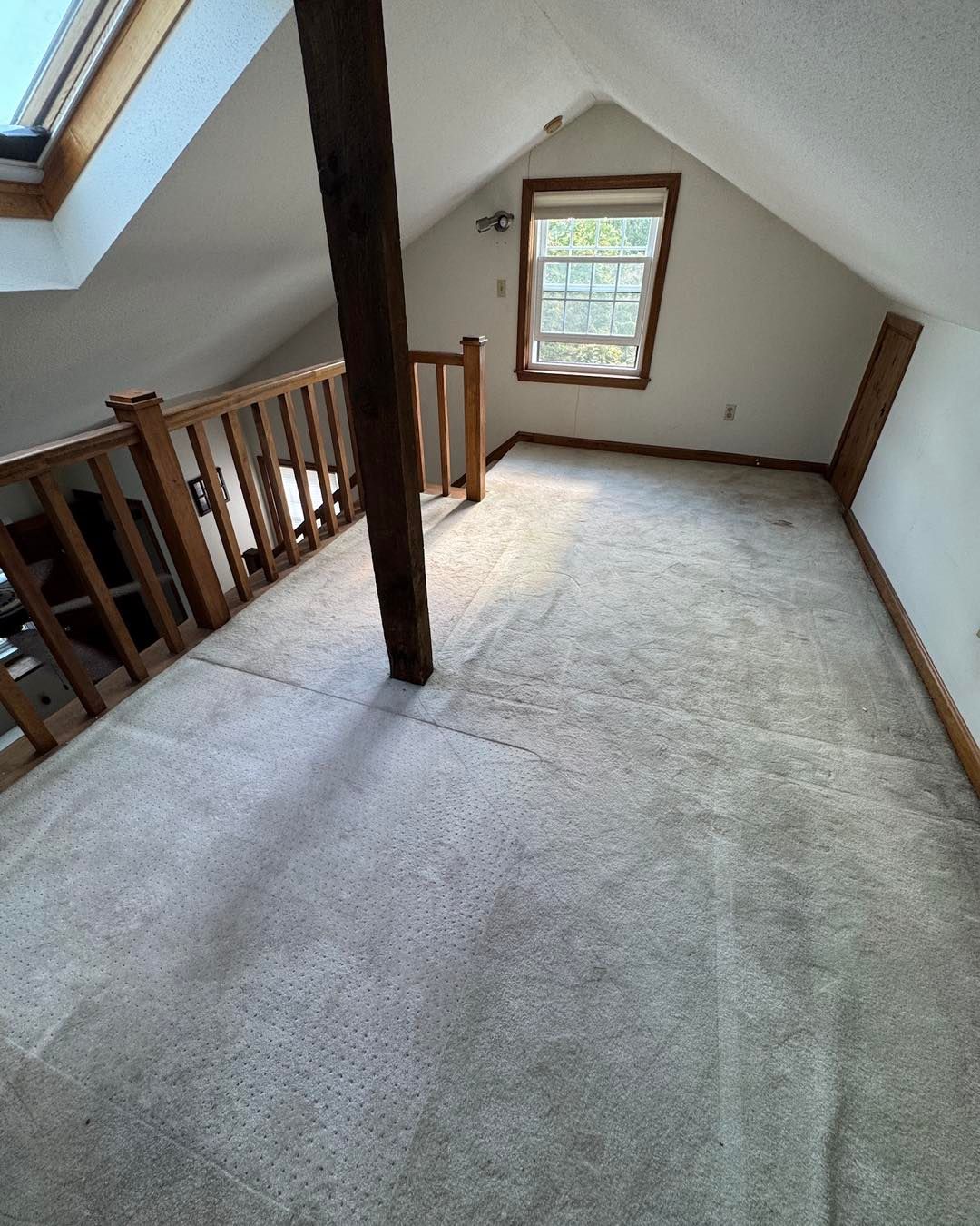Empty carpeted loft with wooden railing, central support post, and a small window.