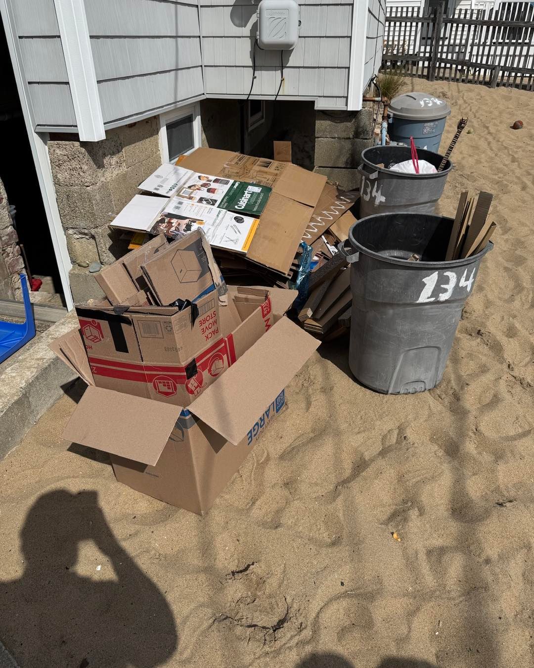 Cardboard boxes and a gray trash can piled outside a house by a basement window.