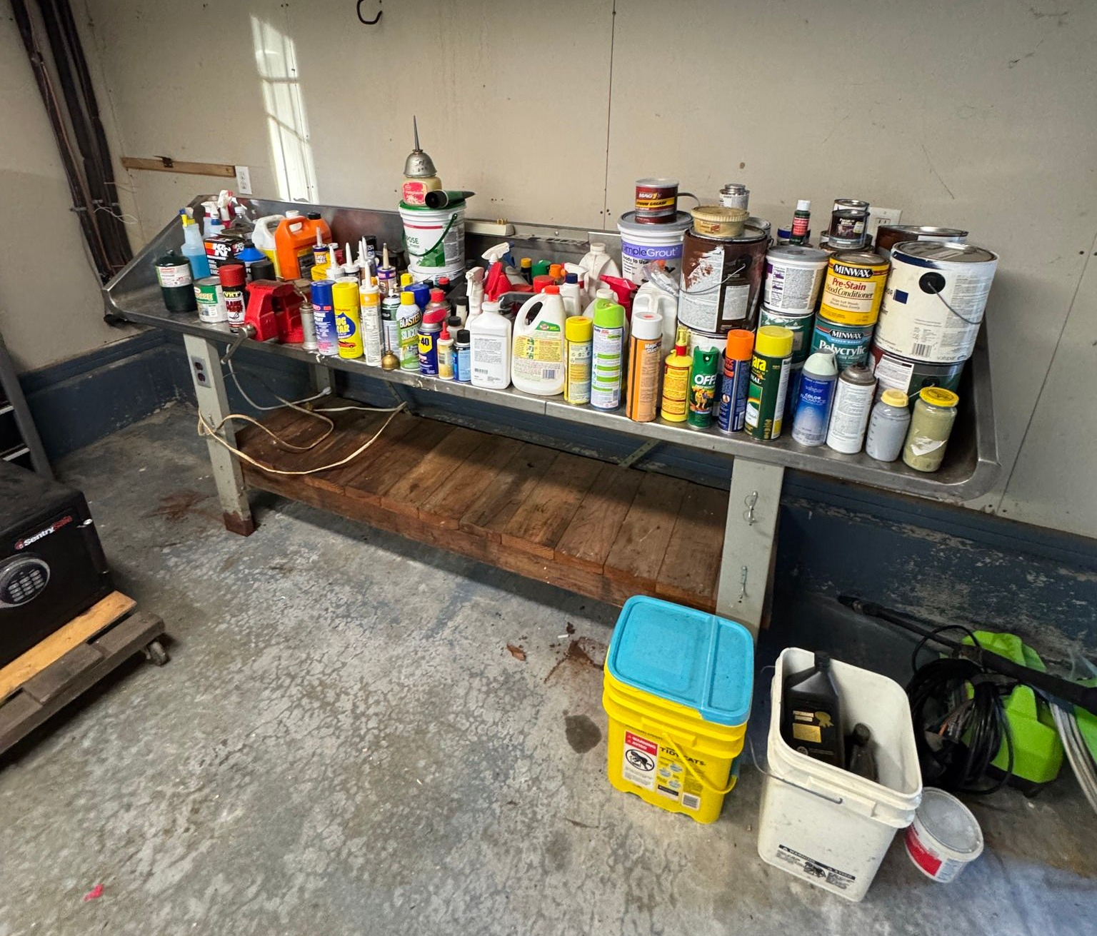 Workbench with many spray cans and chemical containers in a garage, plus buckets and tools underneath.