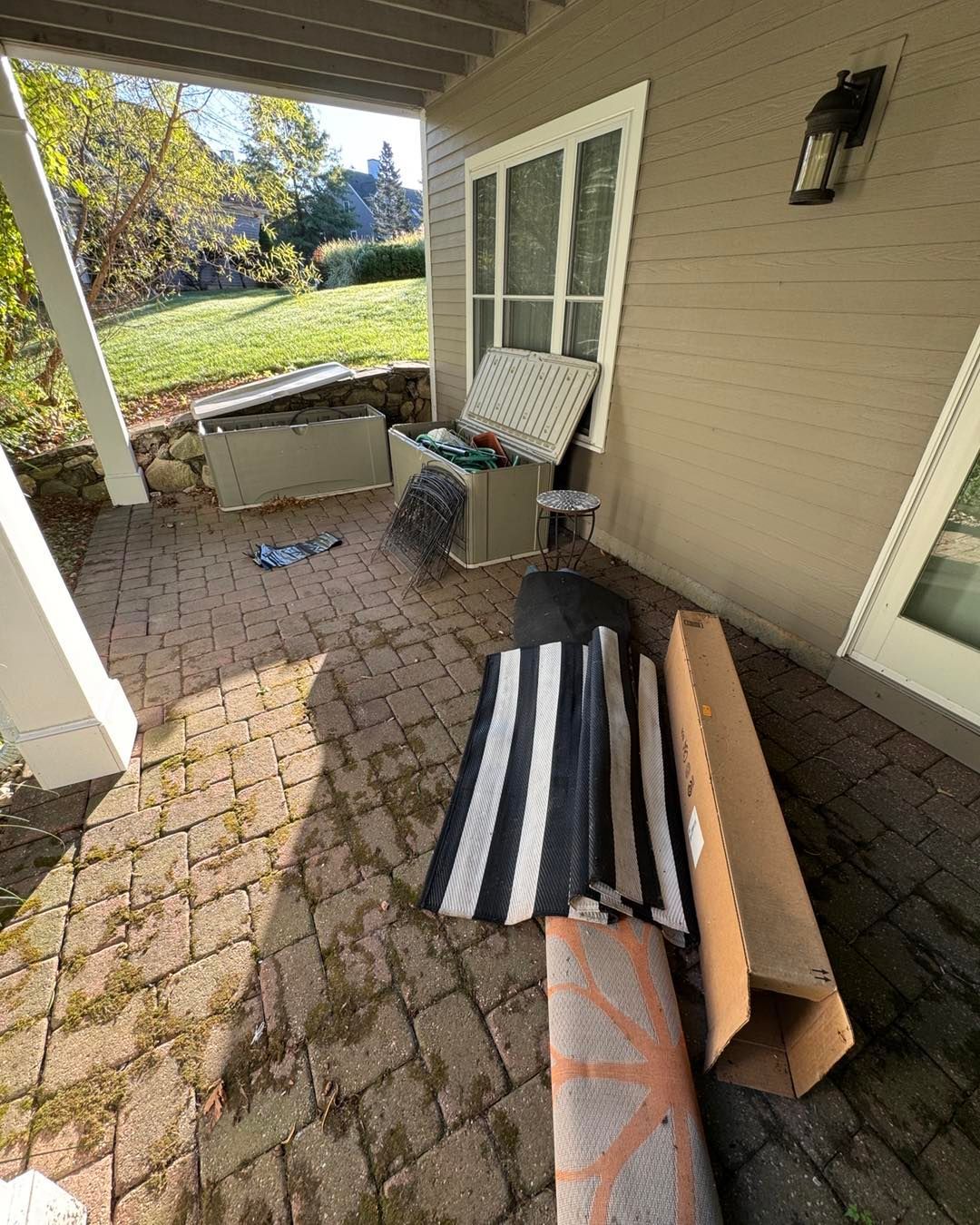Covered patio with folded striped outdoor rug, cardboard, and chair beside a house wall and windows