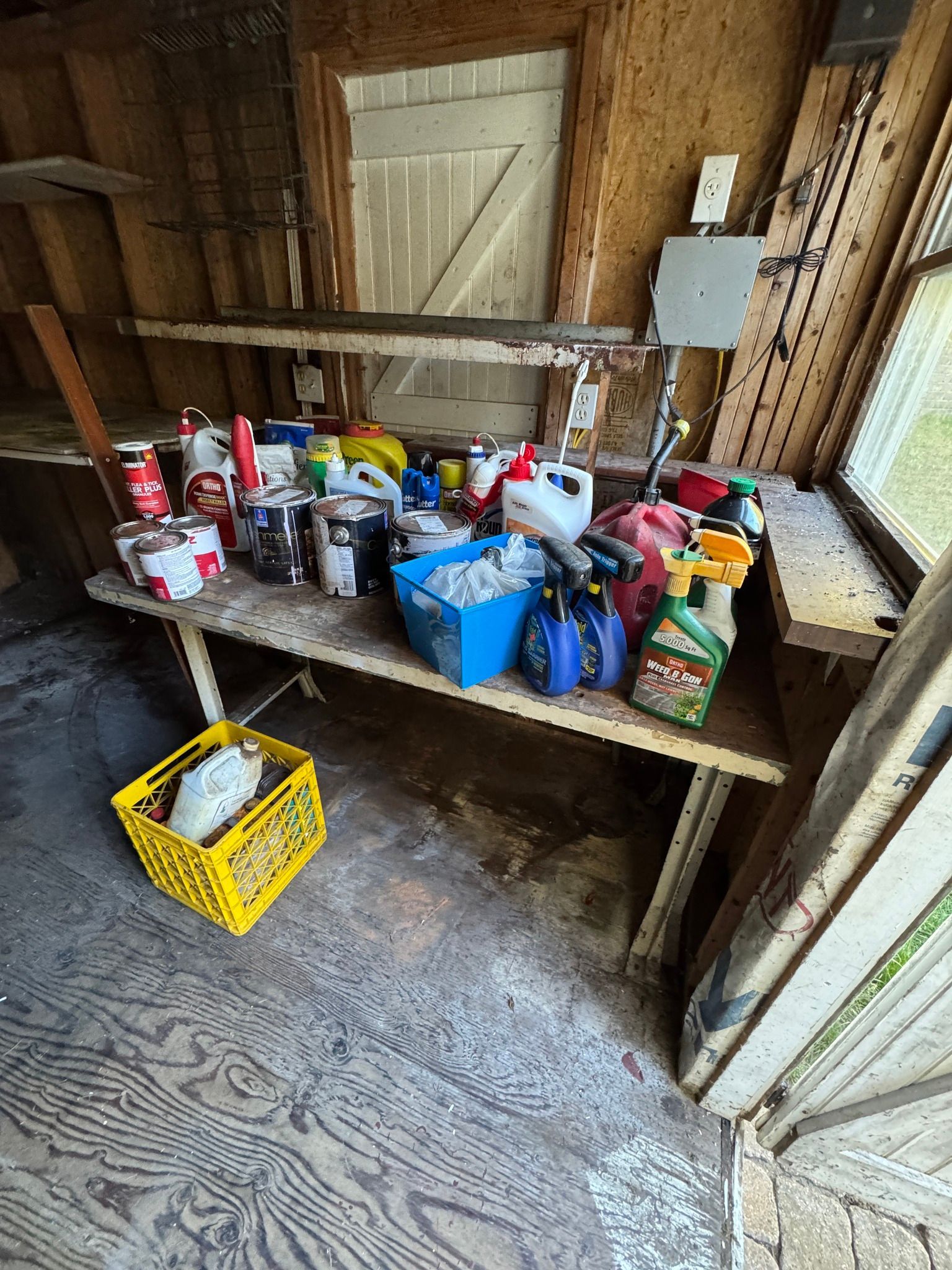 Sheltered workbench cluttered with cleaning supplies, paint cans, and tools in a wooden shed