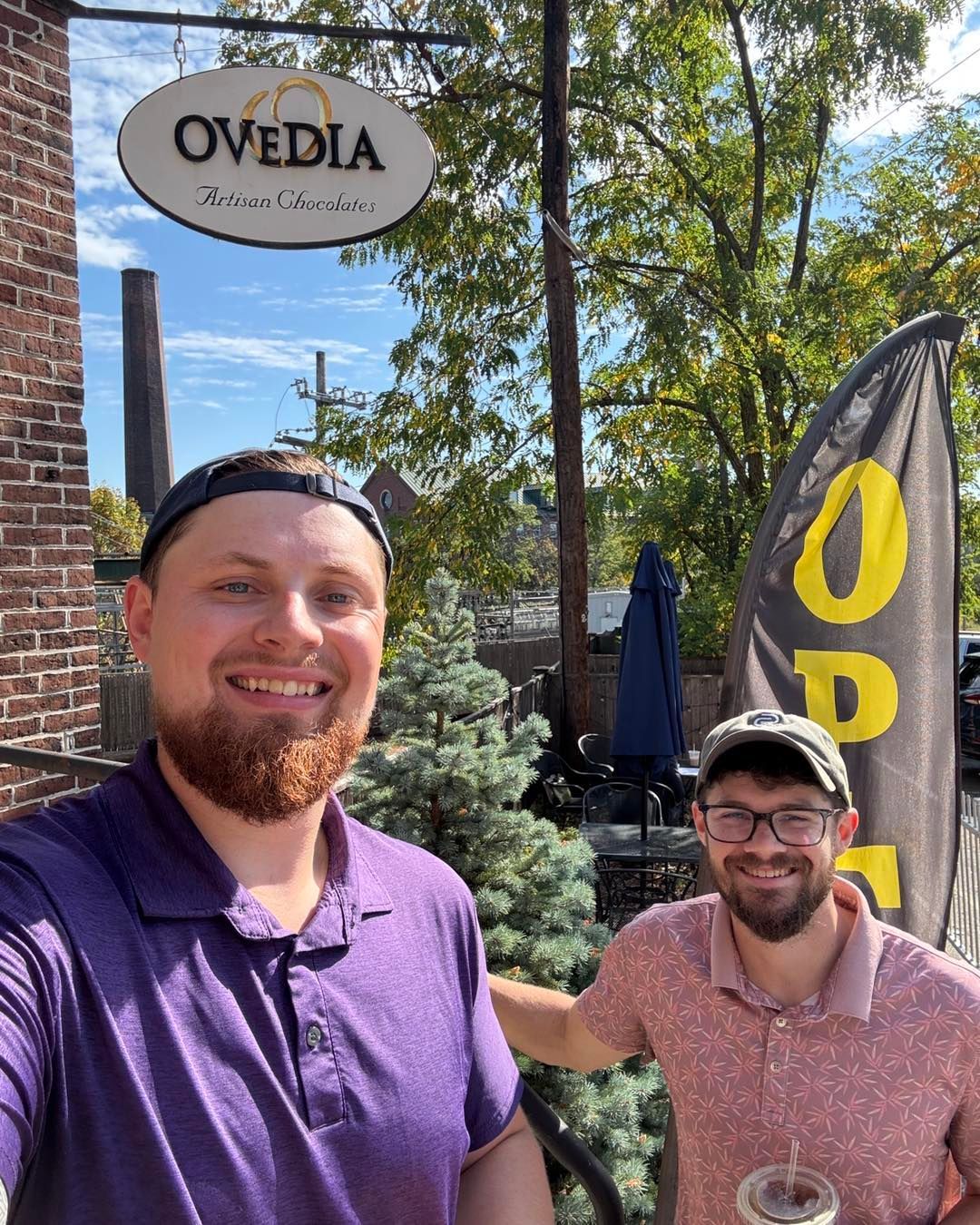 Two smiling men outdoors in front of a sign reading “Ovedia,” one in a purple shirt and one in a pink shirt.
