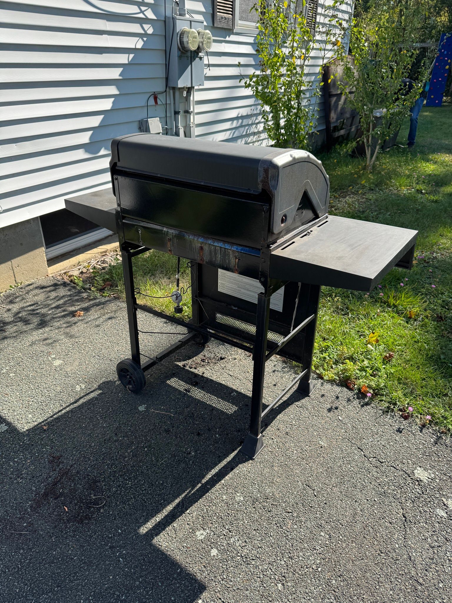 Black outdoor grill with side shelf on a gravel patio beside a house and grass