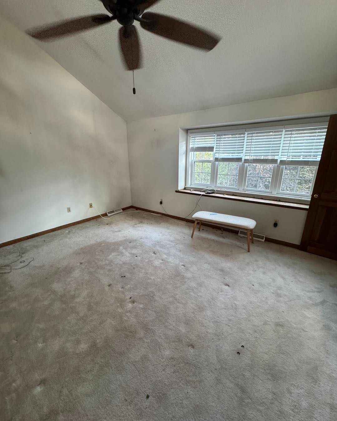 Empty carpeted room with ceiling fan, large window, and white bench near the wall