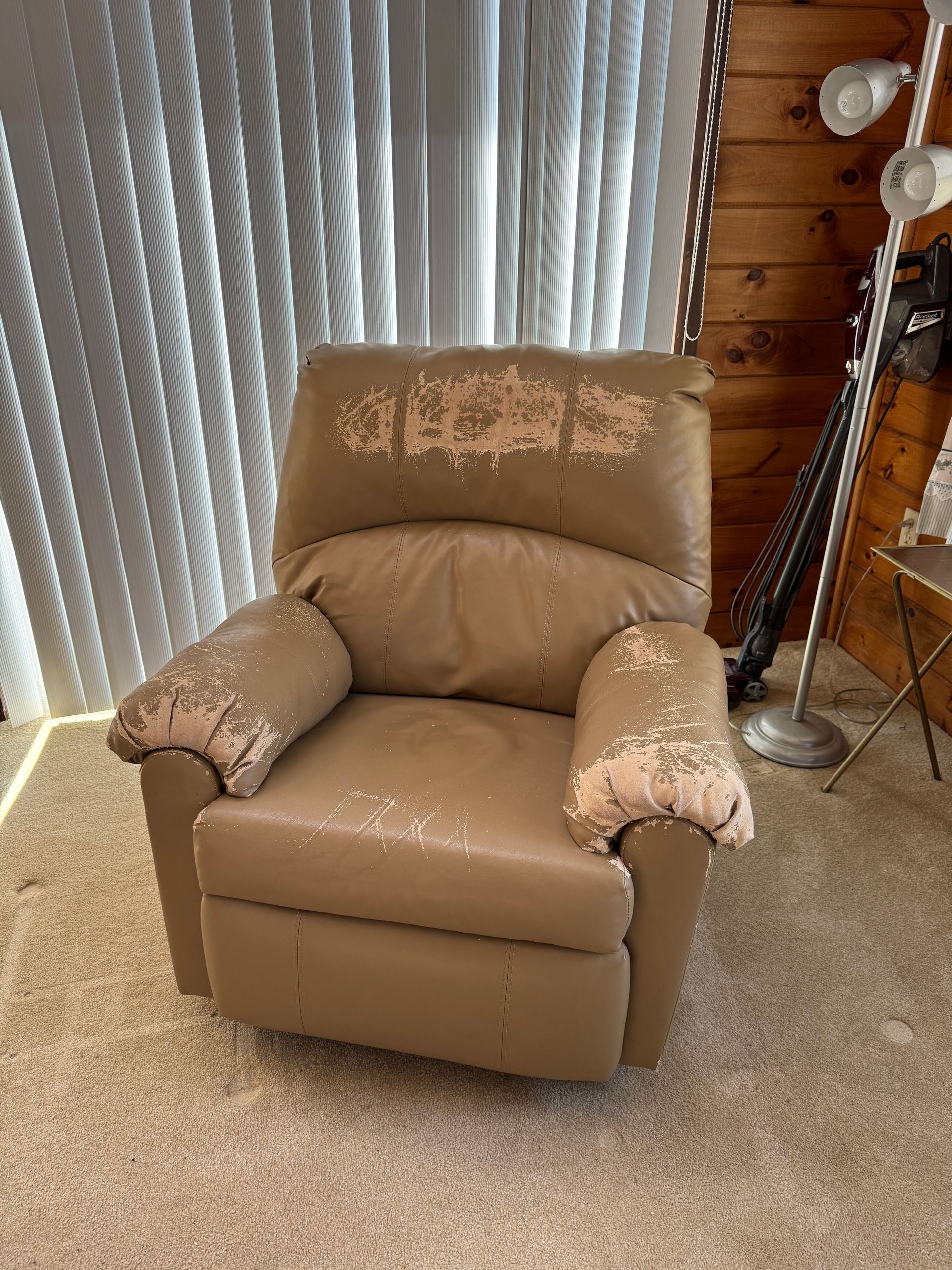 Tan leather recliner chair with floral-patterned fabric on arms and back, in a carpeted room by blinds and a guitar.