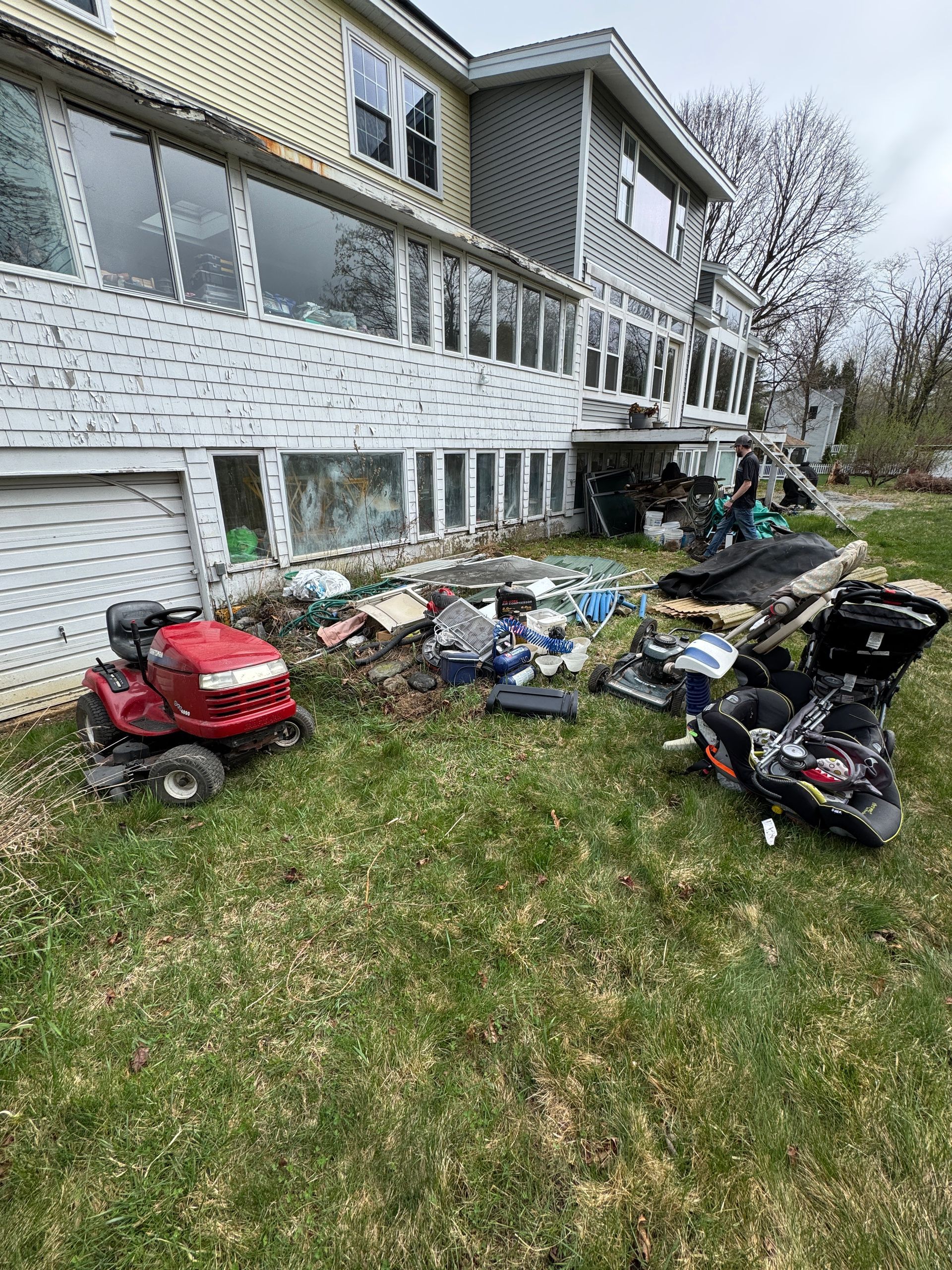 A house exterior with yard tools and lawn equipment scattered across the grass under a balcony.
