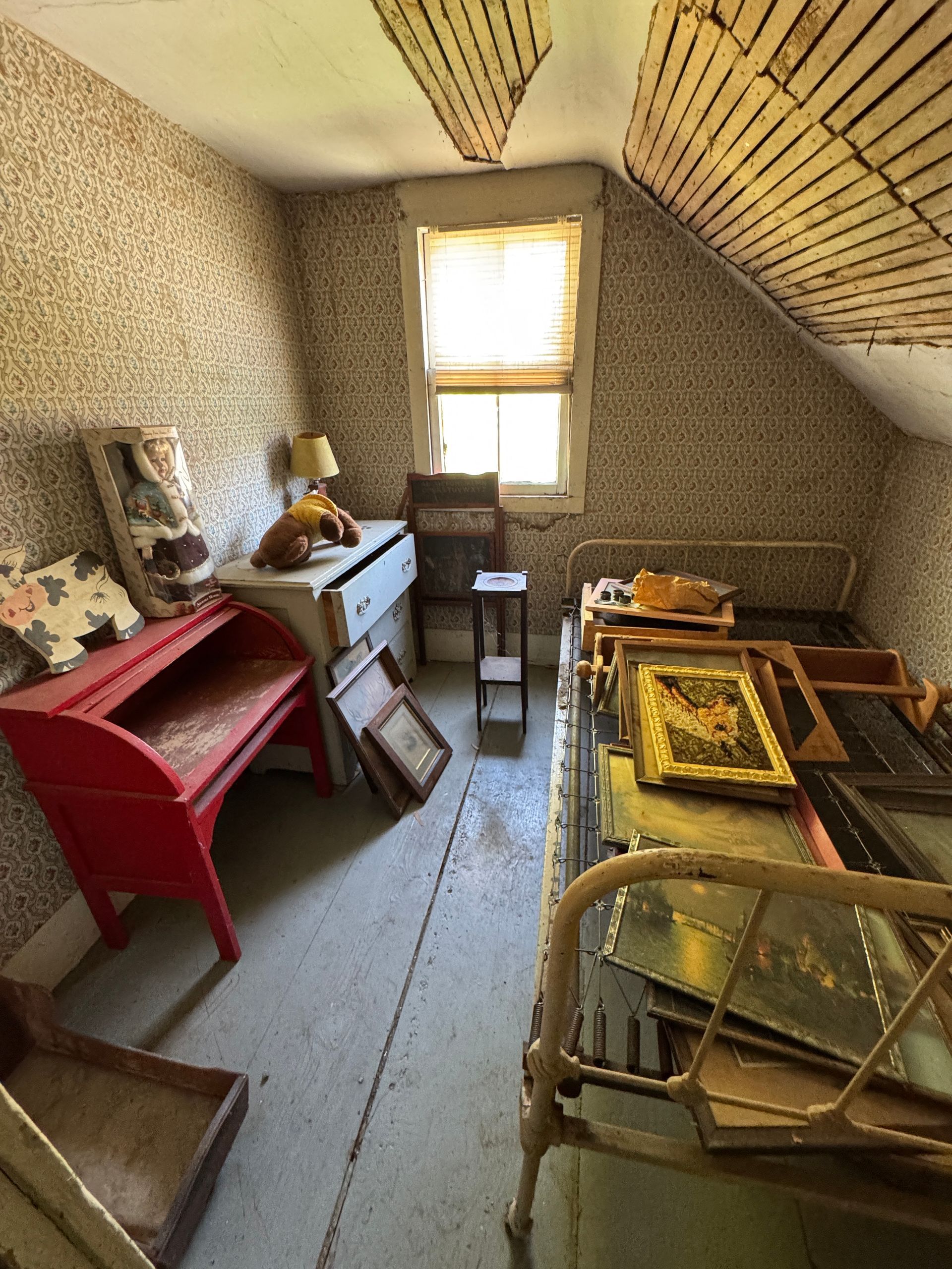 Cluttered attic room with old furniture, red table, framed art, and a small window letting in light