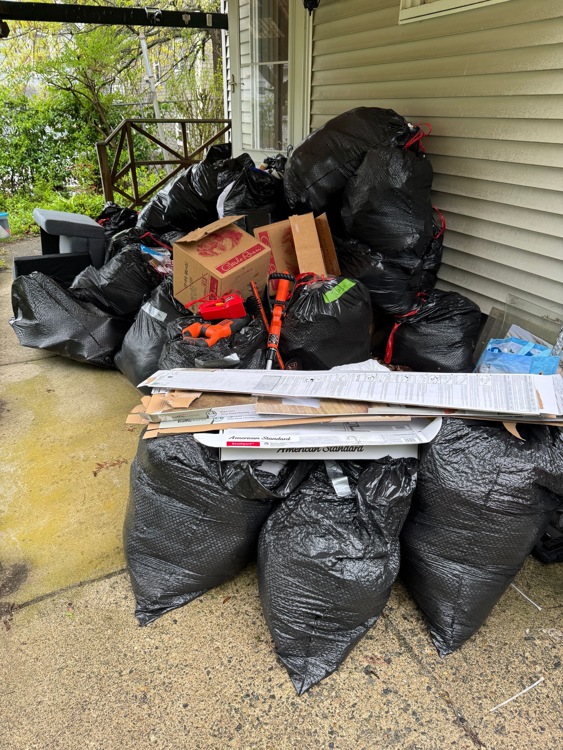 Stacked black trash bags and cardboard boxes piled on a porch beside a house wall.
