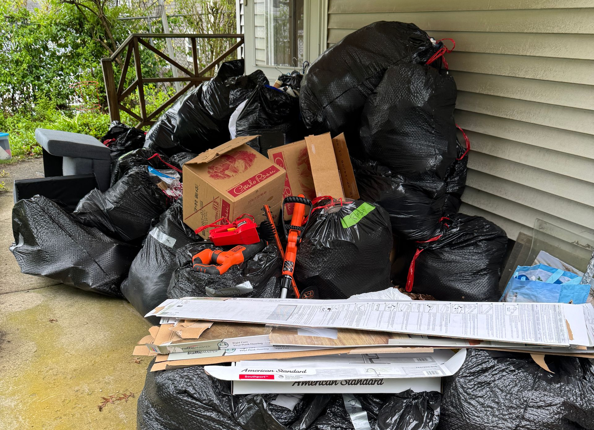 Piles of black trash bags and cardboard boxes stacked on a porch beside house siding.