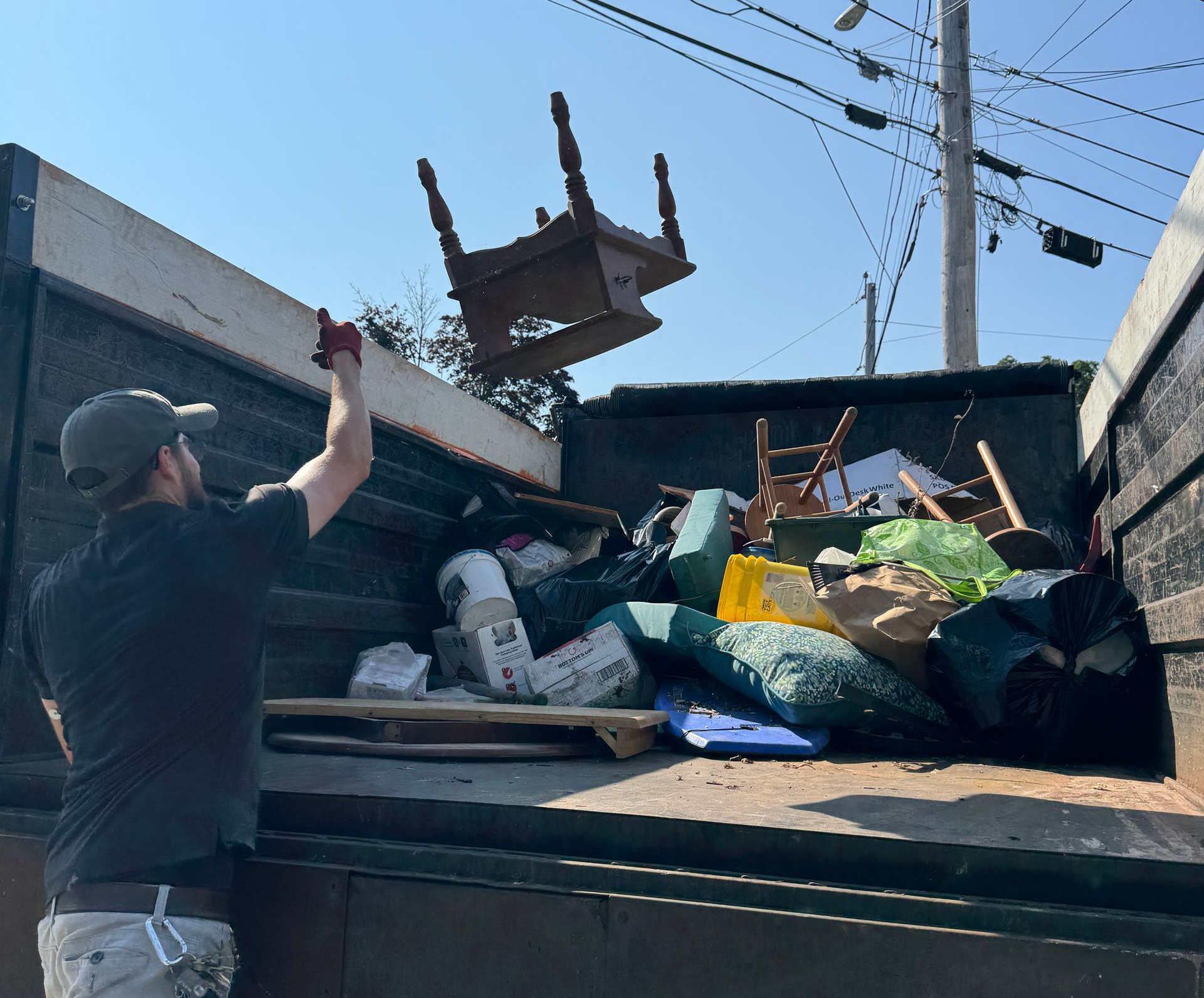Man tossing furniture into a dumpster in bright daylight, with piles of trash and a chair midair