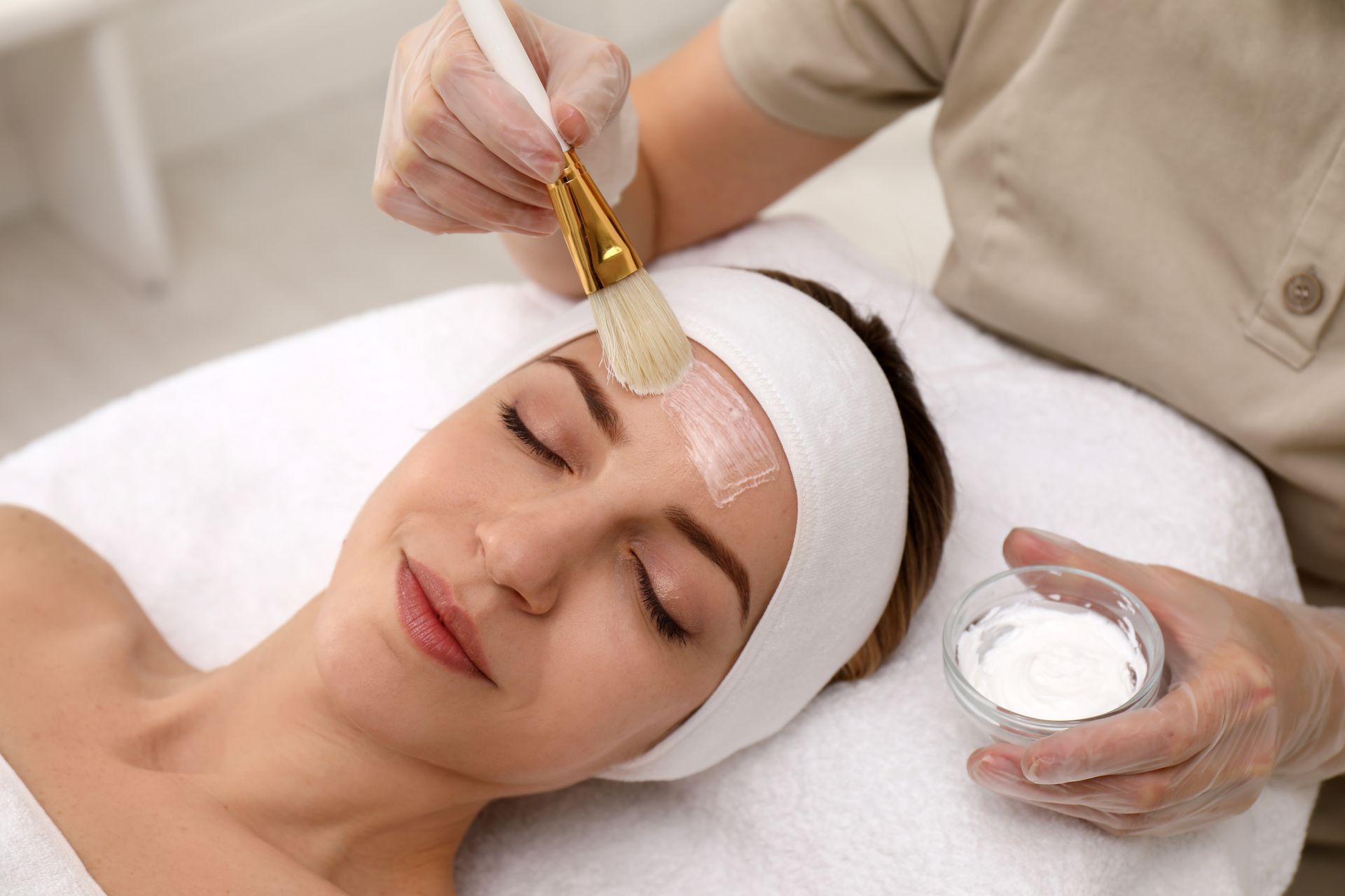 Spa facial treatment with brush applied to a woman’s forehead while a hand holds cream nearby