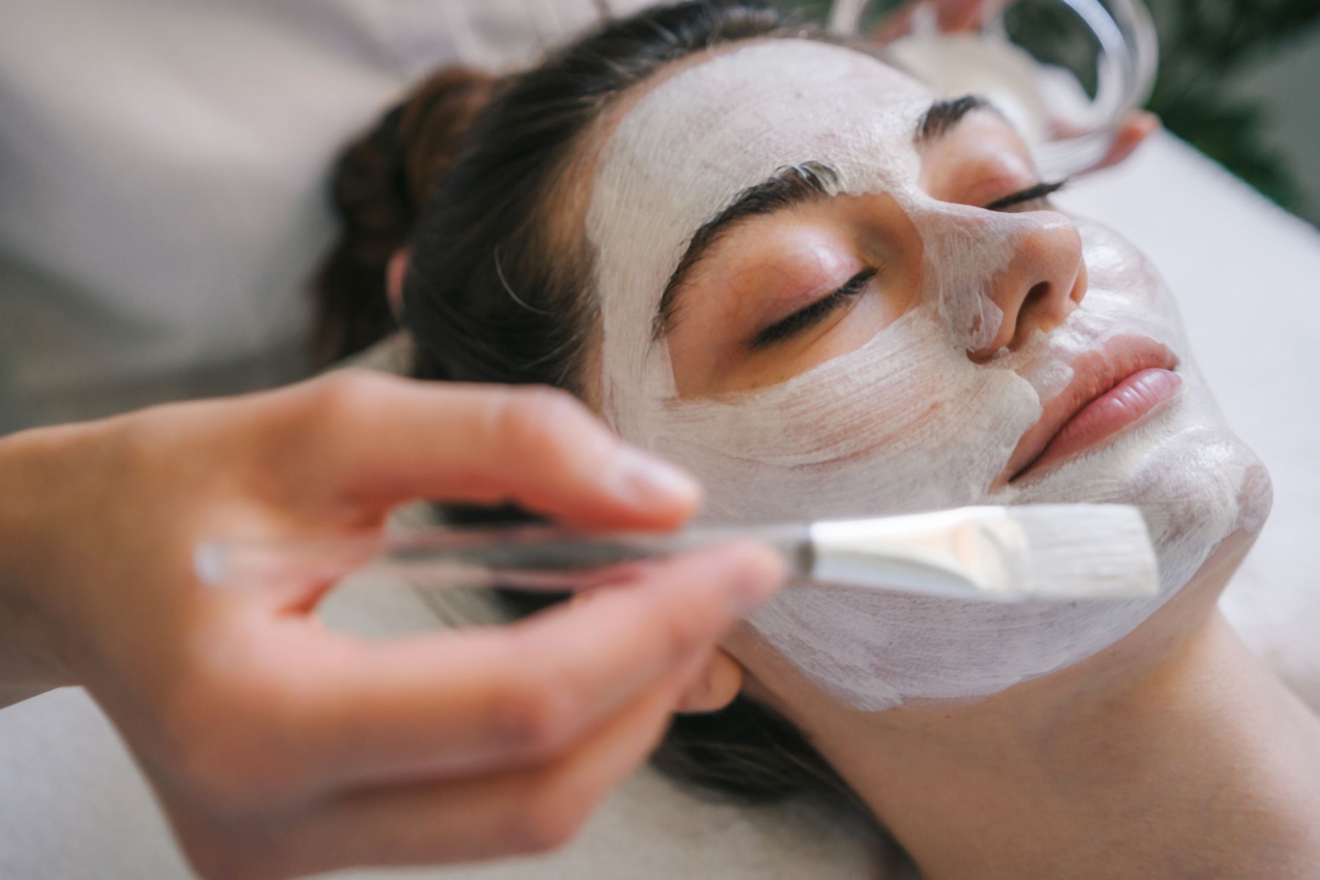 Facial mask being applied with a brush to a person lying down, eyes closed