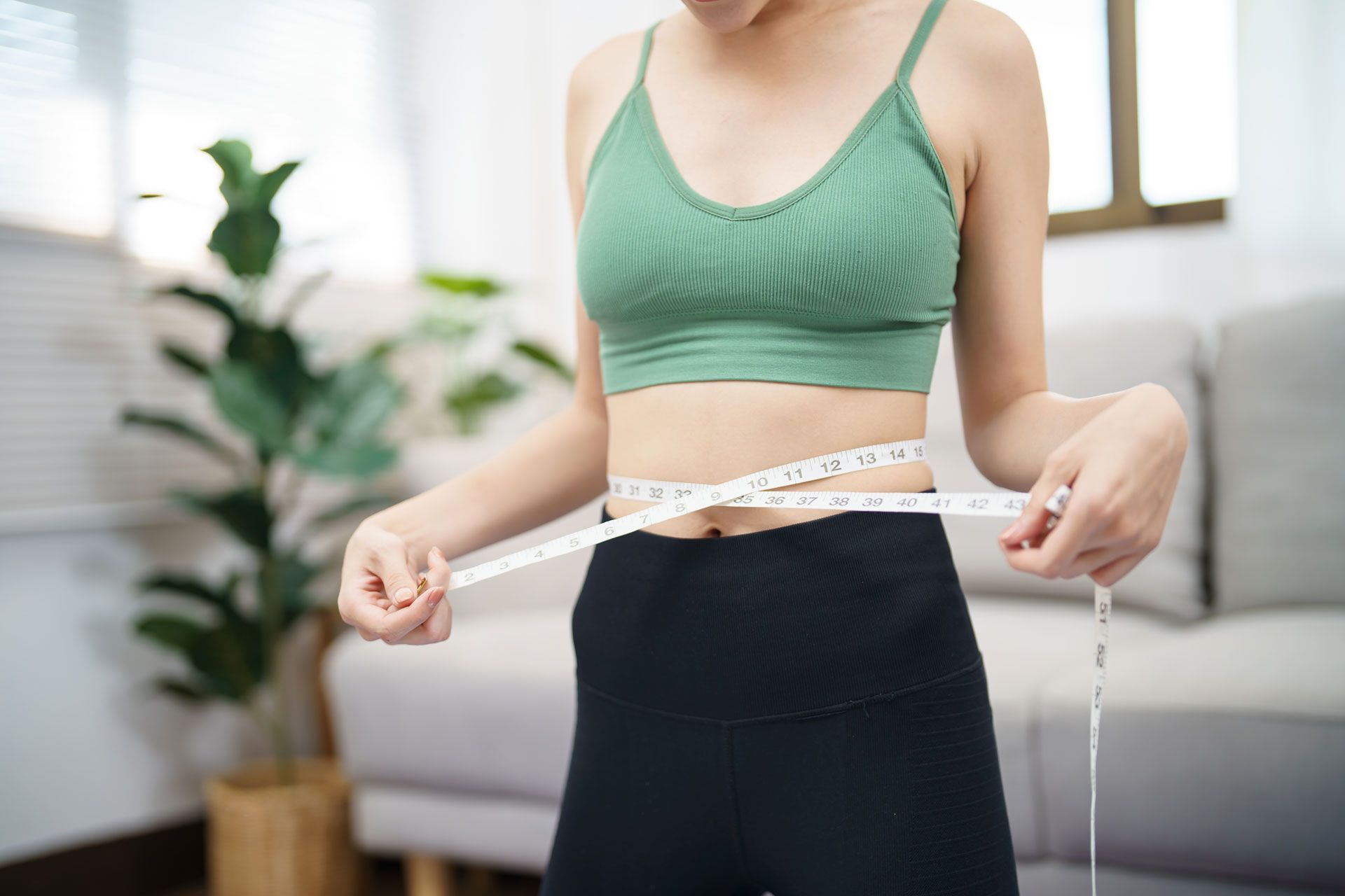 Person measuring waist with a tape measure in a bright room, wearing a green sports bra and black leggings