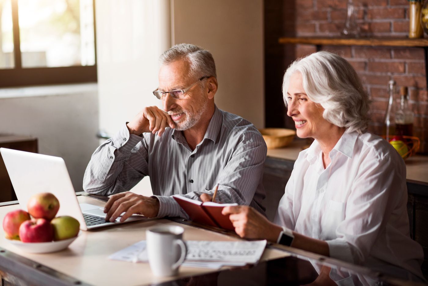 Two people reviewing documents at a table with a laptop, coffee, and fruit by a bright window
