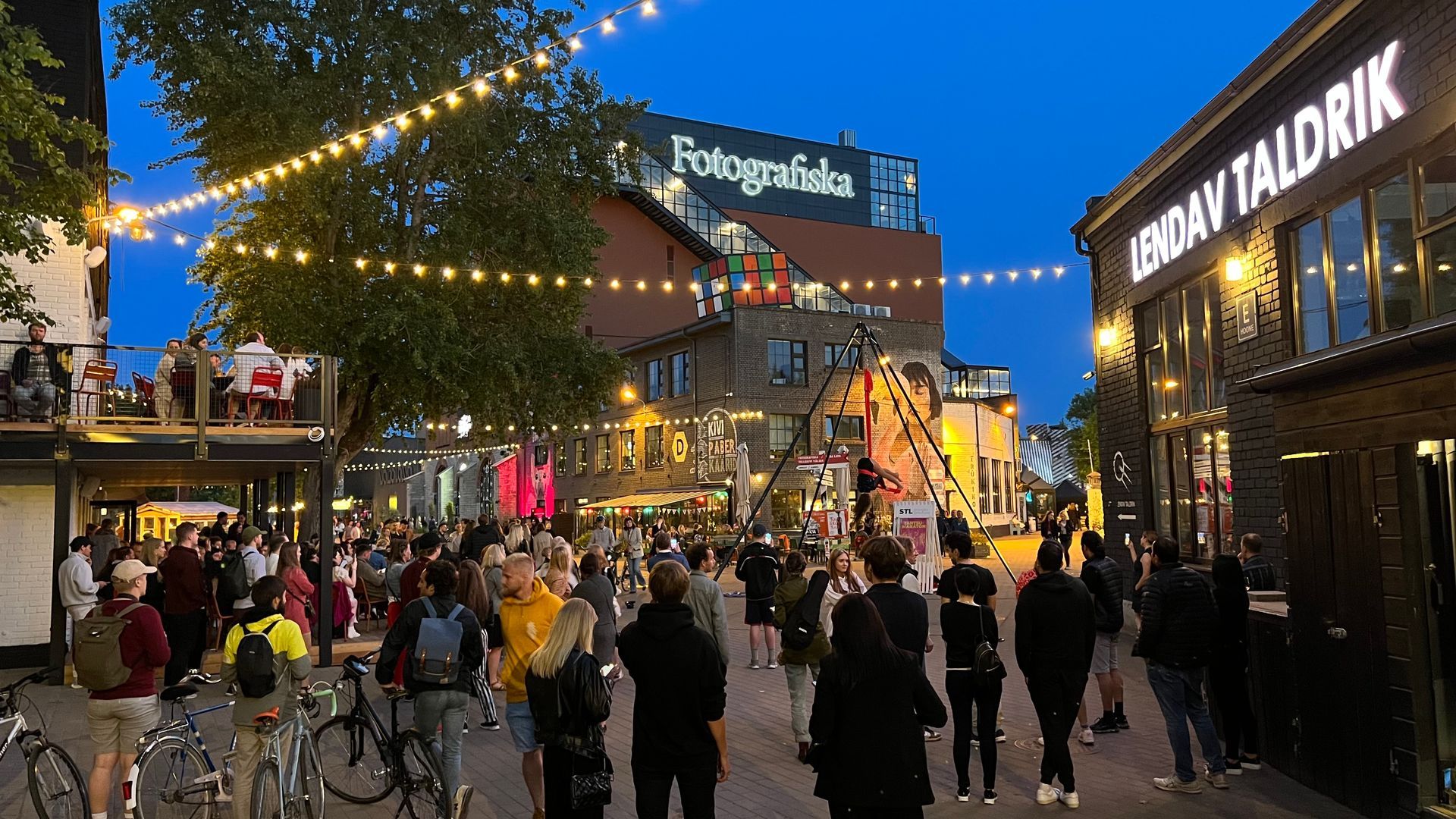 Busy pedestrian street at dusk with string lights, storefronts, and crowds walking past restaurants and shops. One of them being Lendav Taldrik.