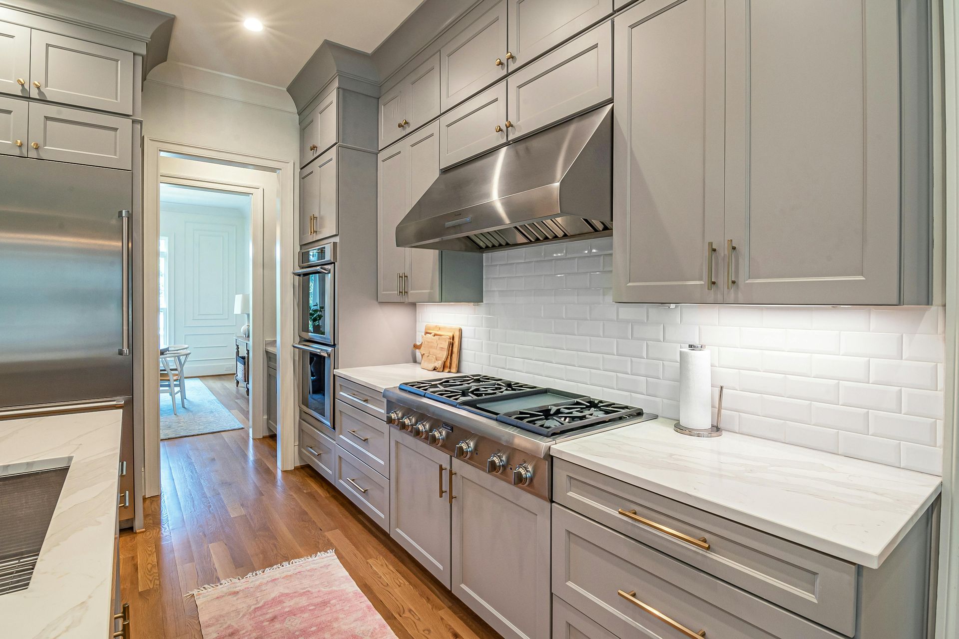 A modern kitchen featuring grey cabinets, a stainless steel hood, gas range, white subway tile backsplash, and wood floors.