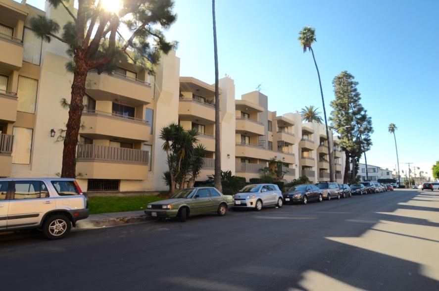Row of beige apartment buildings, cars parked along the street, palm trees, bright sunlight.