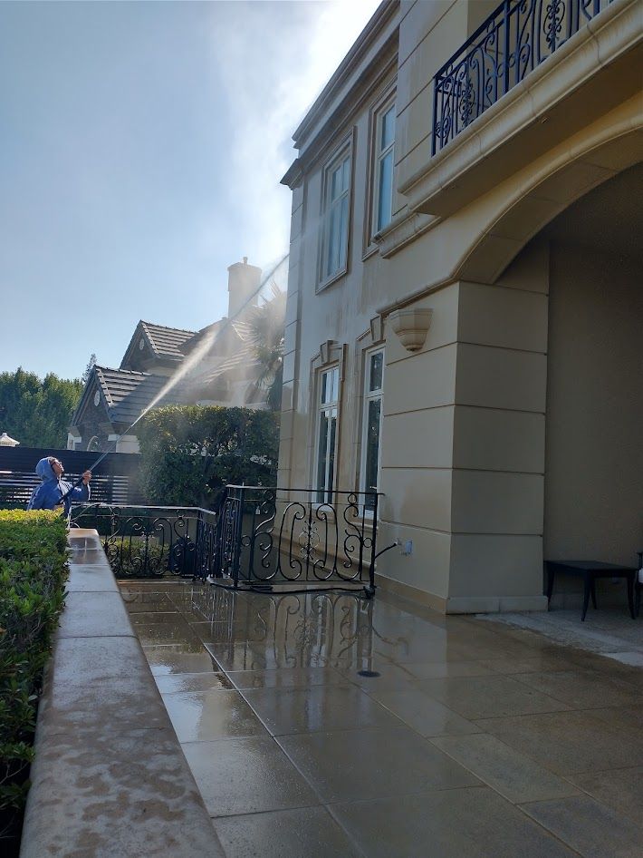 A person power washing the exterior walls of a multi-story, beige-colored stucco home on a sunny day.