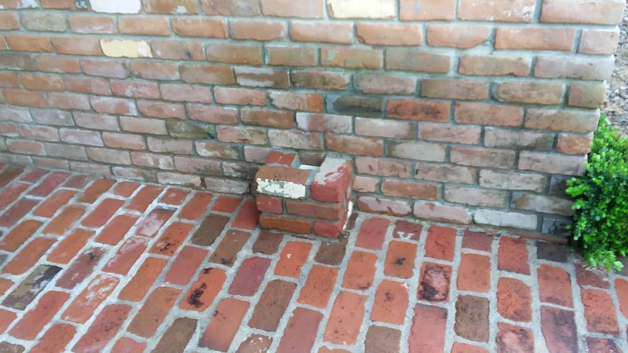 A small, red brick pillar sits against a textured brown brick wall on a brick patio.