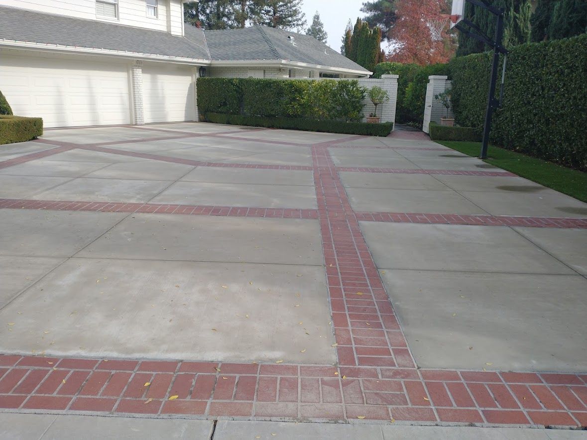 A concrete driveway with a grid pattern of red bricks, set in front of a white house with a landscaped hedge and garage.