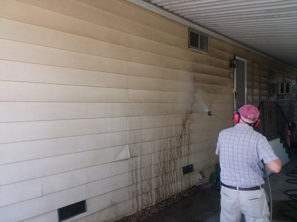 A person wearing protective earmuffs uses a power washer to clean dirt from the light-colored siding of a home.