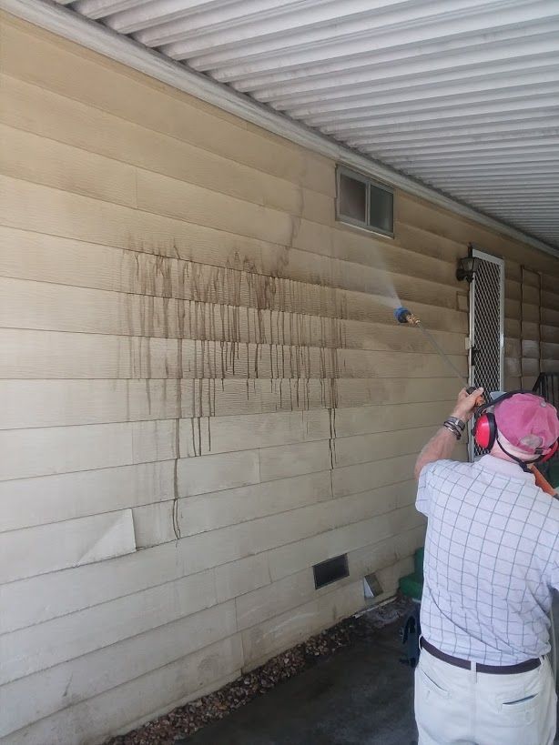 A person wearing ear protection uses a pressure washer to clean vertical siding on a house wall.