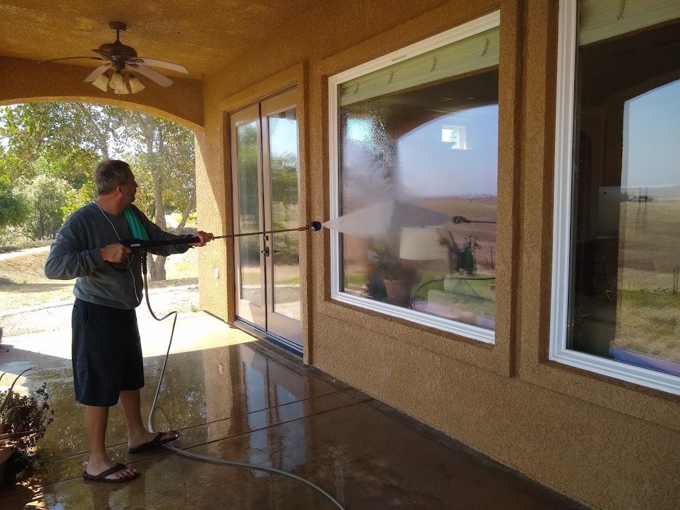 A person standing on a wet patio sprays water from a pressure washer onto the exterior of a home's large glass window.