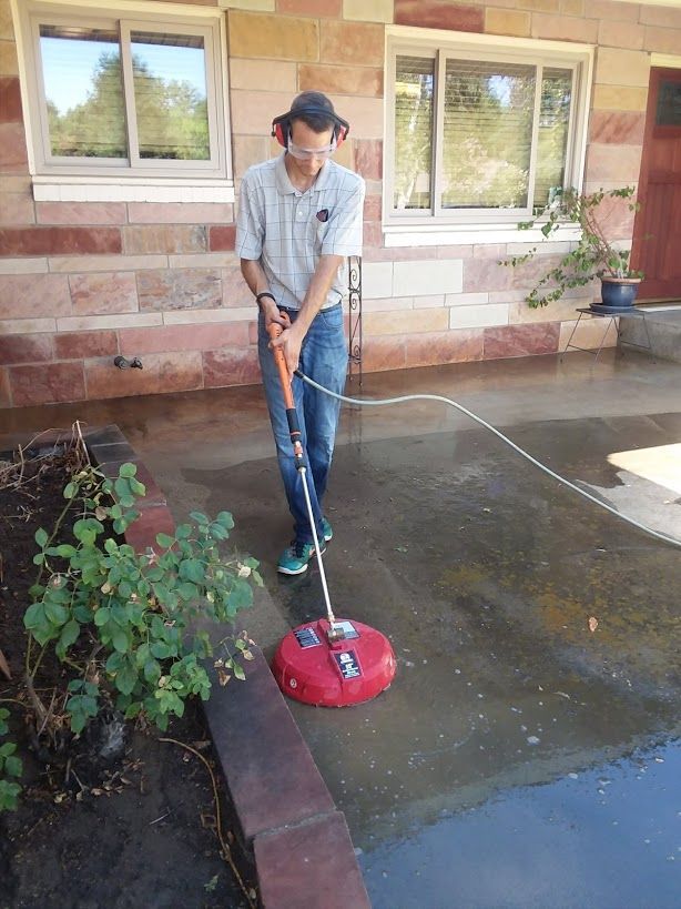 A person wears safety glasses and ear protection while using a red surface cleaner on a wet patio.