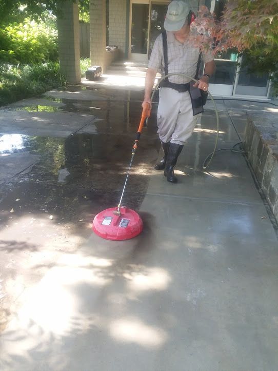 A worker in protective gear uses a red circular pressure washer on a concrete walkway in an outdoor residential setting.