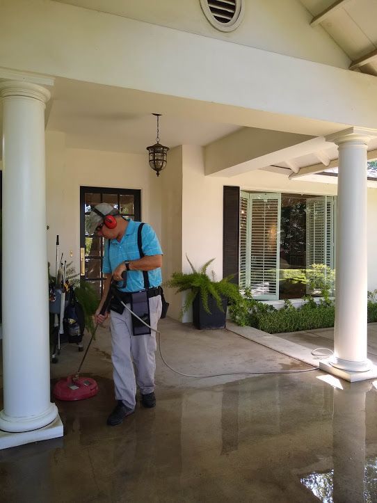 A worker in a blue shirt and ear protection uses a surface cleaner on a wet, covered patio area in front of a house.