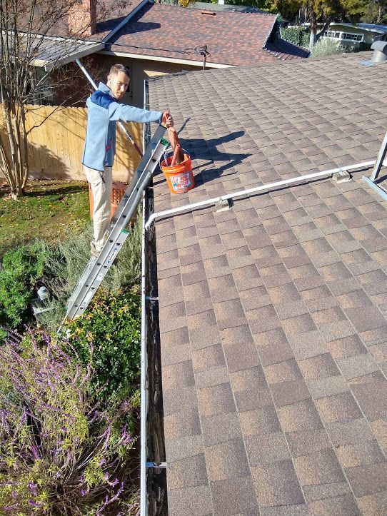 A person in a blue jacket stands on a ladder cleaning debris from a roof gutter into a Home Depot bucket.