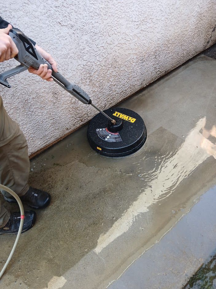 A person uses a DeWalt pressure washer surface cleaner on a concrete patio next to a textured wall.