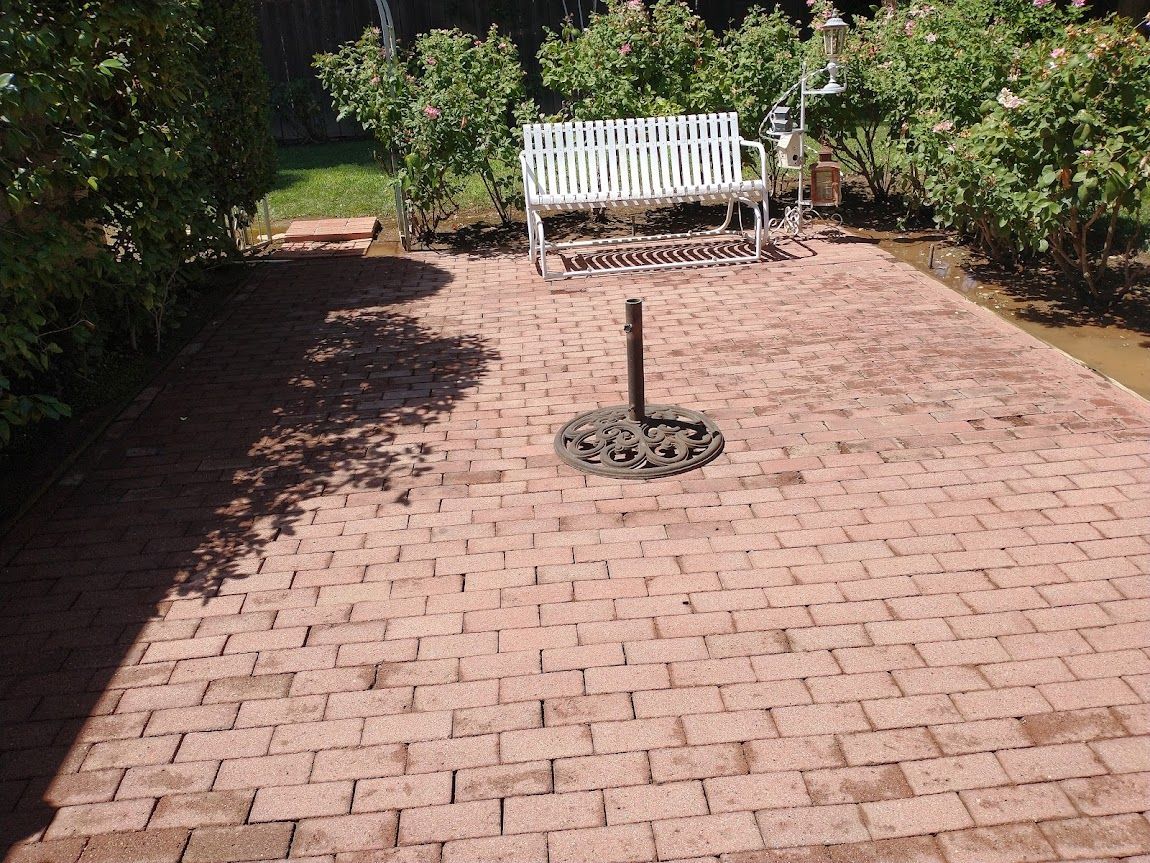 A red brick patio with a central metal pole, a white garden bench, and surrounding bushes in a backyard.