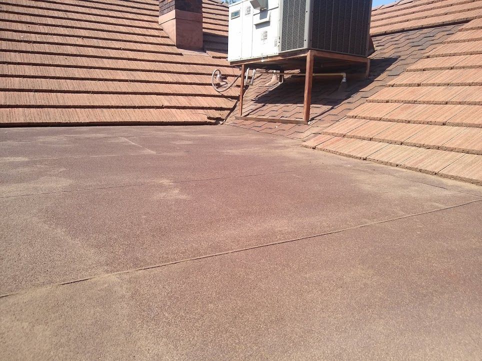 An HVAC unit mounted on a metal stand atop a flat roof section between two sloped, brown tiled roof planes.