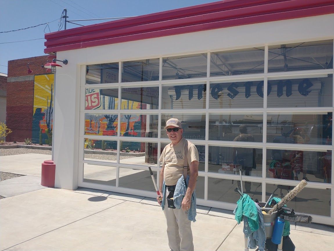 A person stands in front of a building with a large glass garage door and a red roof, with cleaning supplies nearby.