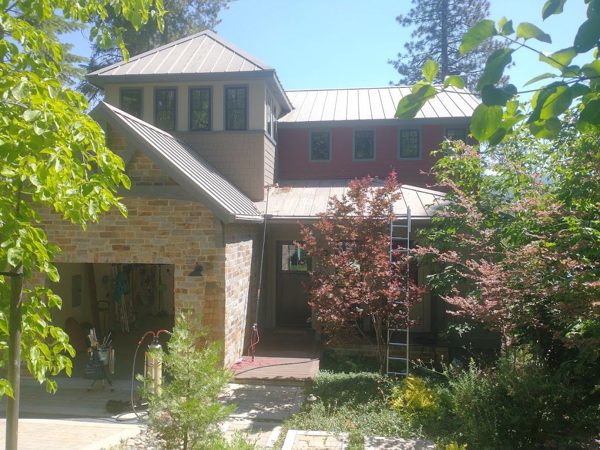 A two-story house with a stone-clad turret, a metal roof, and red siding, partially obscured by lush green trees.