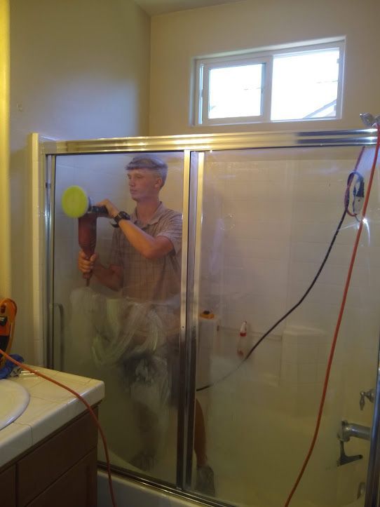A person stands inside a shower stall, using an electric power buffer to clean the glass sliding door.