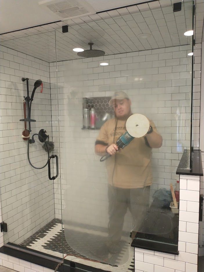 A person stands inside a tiled shower, holding a power buffer to polish the glass door.
