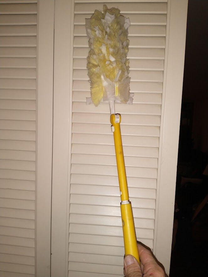 A hand holds a yellow-handled duster with a fluffy white and yellow head in front of white louvered closet doors.