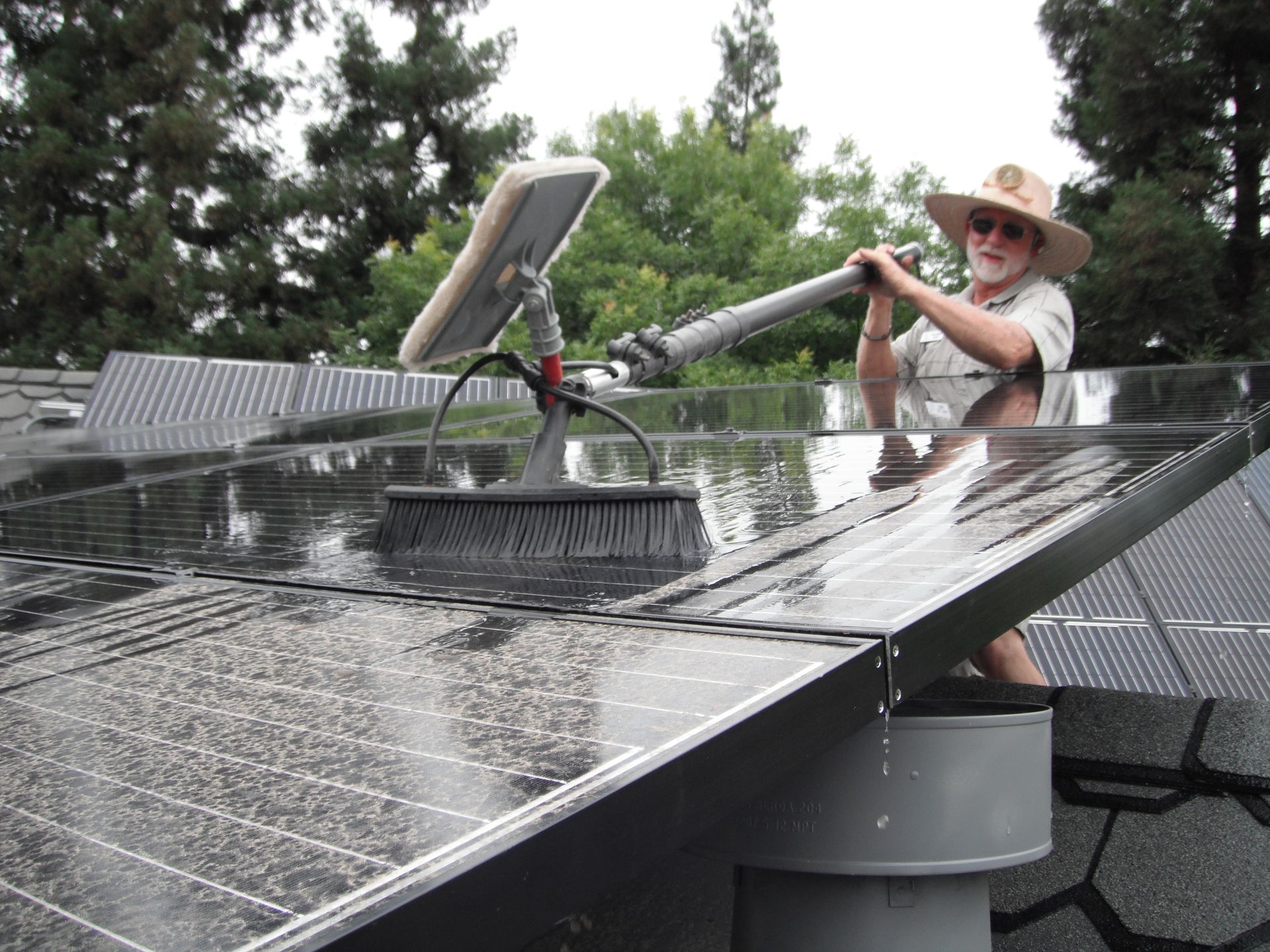 A person wearing a wide-brimmed hat cleans residential solar panels on a roof using a long-handled brush and water.