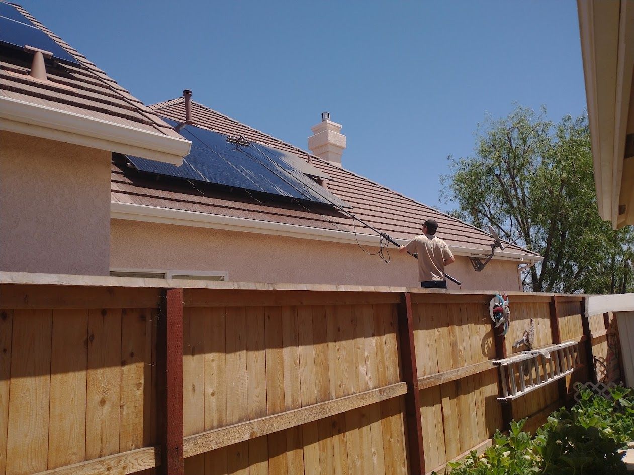 A person on a ladder cleans a tan tiled roof with solar panels, seen over a wooden fence against a clear blue sky.