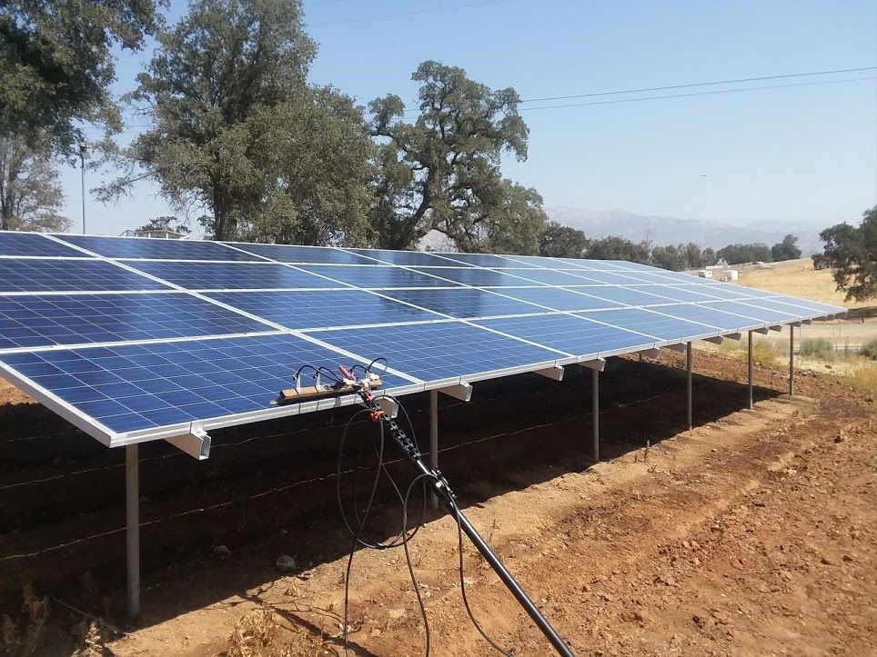 A ground-mounted solar panel array installed in a dry, open field with trees in the background under a blue sky.