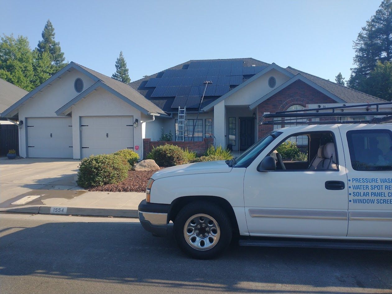 A white work truck parked on a residential street in front of a house with a large solar panel installation on the roof.