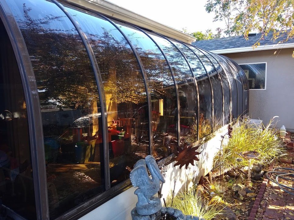 A curved glass solarium attached to the side of a tan house, featuring a dark metal frame and a small garden in front.