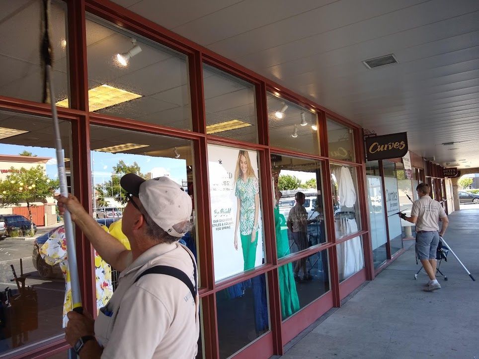 Two people in a storefront entryway use long-handled tools to clean the glass windows of a retail shop.