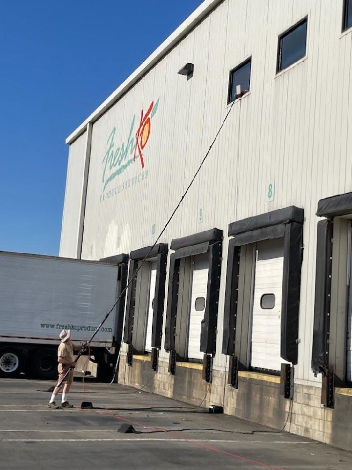 A worker using a long extended pole to reach and clean a high-set window on the side of a large warehouse building.