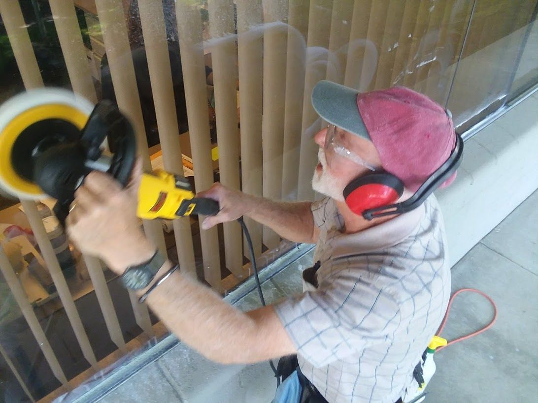 A person wearing a cap, glasses, and earmuffs uses a power buffer tool to polish a large glass window.