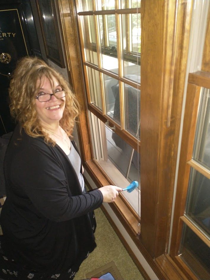 A smiling person wearing glasses and a black cardigan cleans a wooden-framed window with a blue tool.